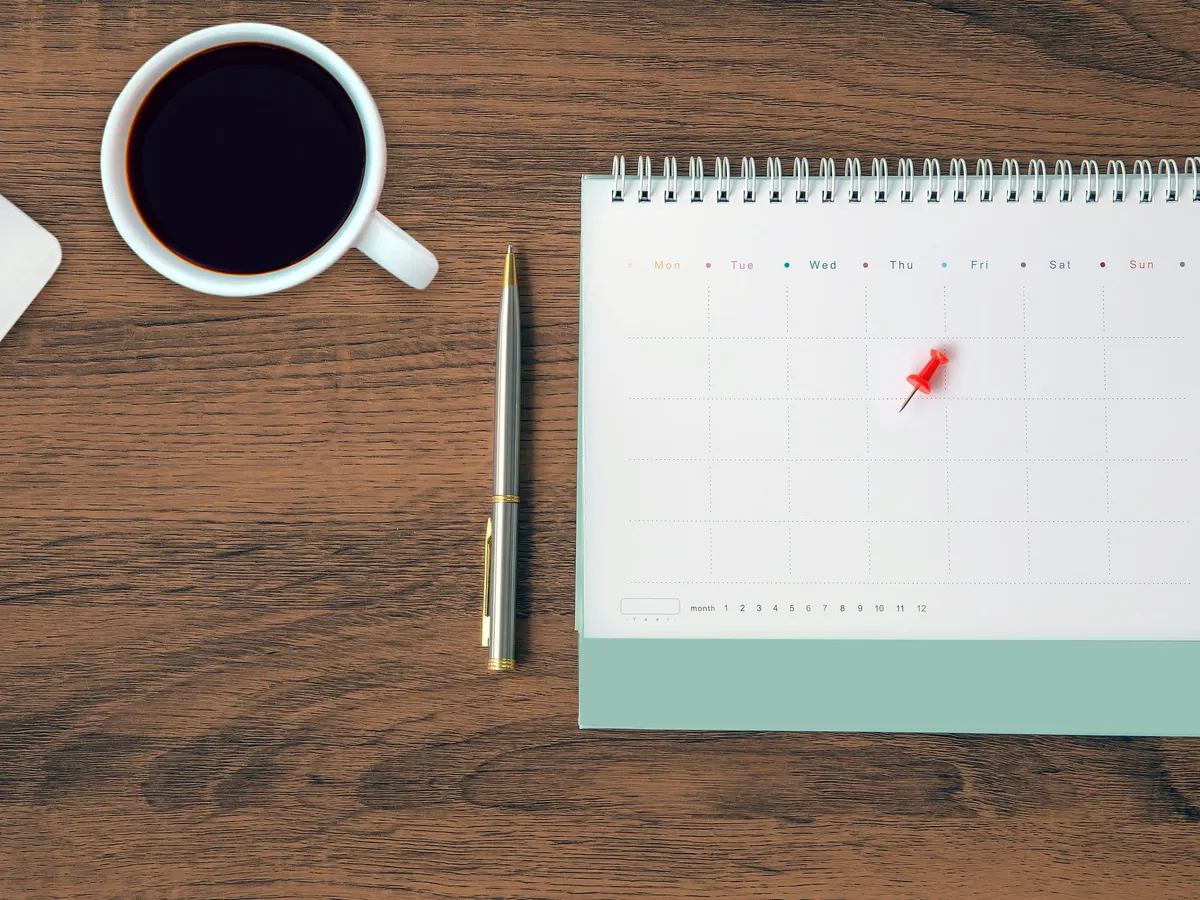 A desk calendar with a red pin, a pen and a cup of coffee on a wooden desk — a stock picture