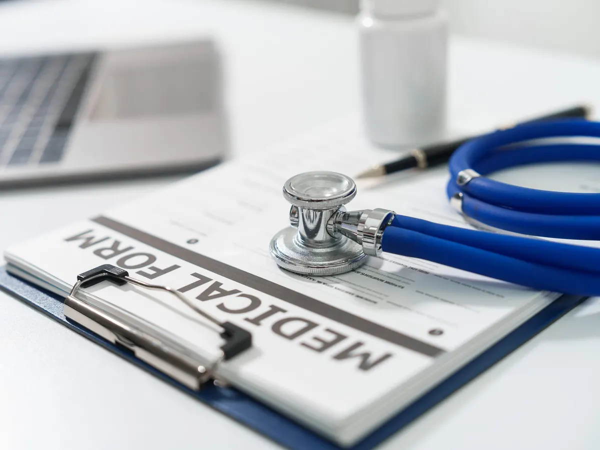 A blue stethoscope and a pen lying on a medical form on a clipboard beside a laptop — a stock picture