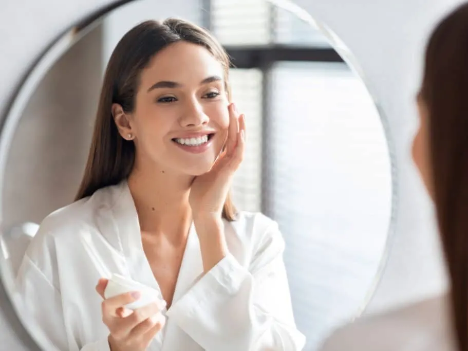 Woman smiling as she is looking in the mirror applying skincare for the winter