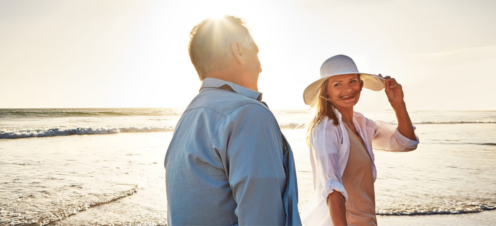 A smiling woman in a sun hat and a man walking along the beach at sunset